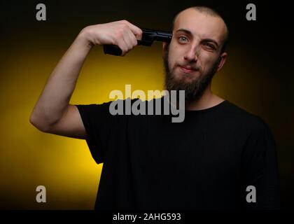 Ritratto di un giovane uomo che punta una pistola alla sua testa. Colpo contro scuro e sfondo giallo Foto Stock