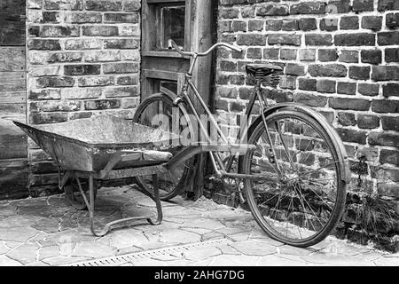 Una vecchia bicicletta appoggiata contro un rosso un muro di mattoni, accanto a una carriola arrugginito lasciato all'entrata dell'edificio. Foto Stock