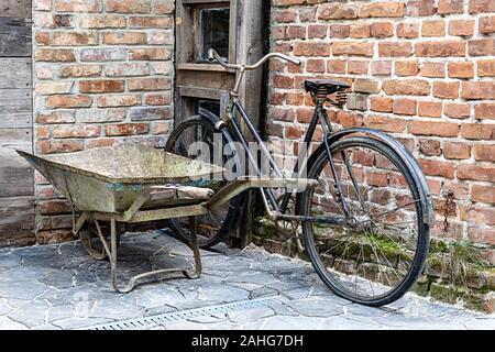 Una vecchia bicicletta appoggiata contro un rosso un muro di mattoni, accanto a una carriola arrugginito lasciato all'entrata dell'edificio. Foto Stock