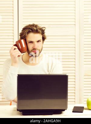 Freelance lavoro a casa e la pausa pranzo concetto. Uomo Barbuto con tazza vicino alla guancia sulla luce sfondo striato. Guy utilizza laptop e detiene un tè o un caffè mug. L'uomo con la faccia concentrata e gadget moderno Foto Stock