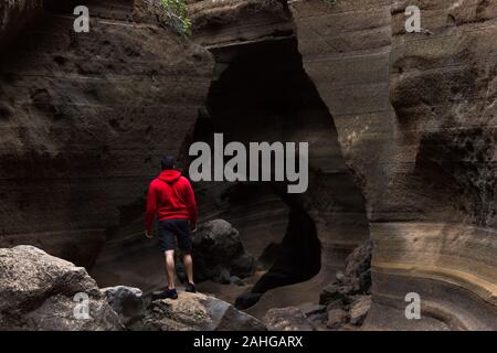 Retro del giovane uomo su felpa con cappuccio rosso permanente sulla cima della roccia marrone rocce formazione canyon in Gran Canaria, Spagna. Lonely avventuriero escursionismo Foto Stock