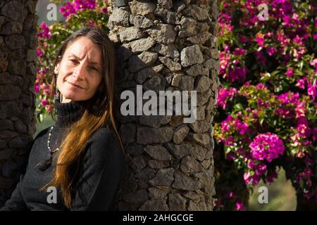 Modello femminile biondo a pelo lungo appoggiato su colonna di pietra con fiori rosa sullo sfondo. Donna sorridente bella che posa nel parco giardino Foto Stock