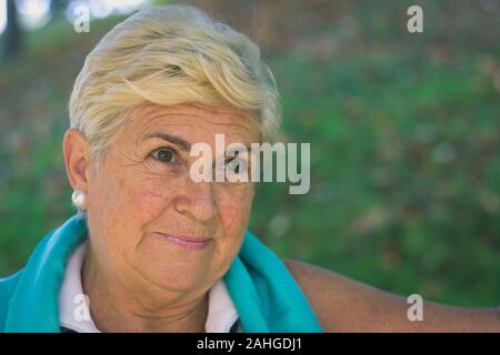 Malinconici elder lady ritratto nel parco. Senior donna bionda sorridente con sguardo ottimista. Primo piano della persona matura con sleeveless camicia bianca Foto Stock