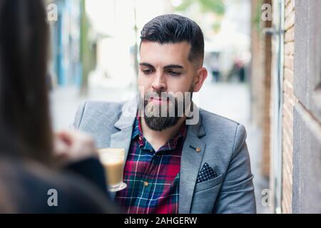 Un bel uomo d'affari che guarda il caffè in vetro sulla terrazza del caffè in città. Modello di hipster a barba intera su blazer grigio deliziato con cappuccino tazza Foto Stock