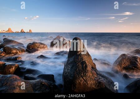 Nel tardo pomeriggio su Aci Trezza e loro cataste di mare con onde che si infrangono sulle rocce in primo piano, Catania, Italia Foto Stock