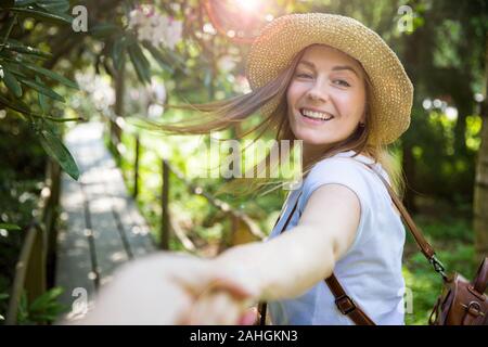 Bella donna in cappello di paglia viaggio nella foresta tropicale, camminando lungo il sentiero di legno, che conduce il suo partner. Turistico con zaino. Seguimi il concetto. Foto Stock
