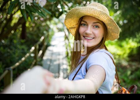 Bella donna in cappello di paglia viaggio nella foresta tropicale, camminando lungo il sentiero di legno, che conduce il suo partner. Turistico con zaino. Seguimi il concetto. Foto Stock