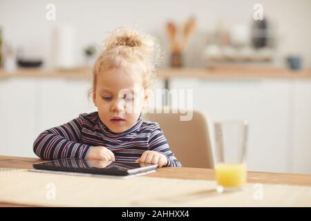 In tonalità calde ritratto di carino bambina con tavoletta digitale mentre è seduto al grande tavolo in accogliente cucina, spazio di copia Foto Stock