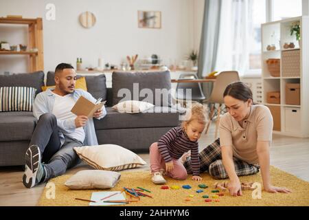 A piena lunghezza Ritratto di felice famiglia moderna giocando con graziosi bambina in accogliente soggiorno interno, spazio di copia Foto Stock