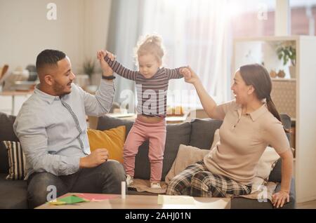 Dai toni caldi ritratto di felice famiglia moderna giocando con incantevole piccola figlia in casa accogliente interno