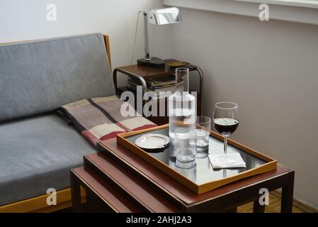 Home interno con still life - un bicchiere di vino rosso, due bicchieri e caraffa, posacenere sul vassoio sul tavolo, angolo di camera con mobili e lampada Foto Stock