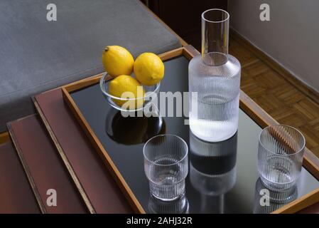 Tre freschi limoni giallo in una ciotola di vetro, due bicchieri e la caraffa con acqua sul vassoio moderno su tavola, still life Foto Stock