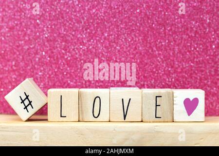 La parola amore su cubi di legno con cuore rosa e hashtag, close-up vicino a glitter sfondo rosa Foto Stock