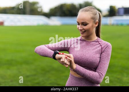 Sport e tecnologia. bellissima giovane donna whiteskinned con coda di cavallo alla esecuzione di stadium di fronte di allenamento utilizza una scattante Smart clock sul suo braccio per Foto Stock