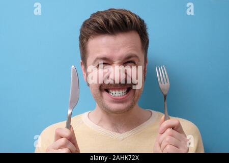 Crazy, affamati uomo con i baffi tiene un coltello con una forcella nelle sue mani e sorrisi. Sto morendo di fame concetto. Foto Stock