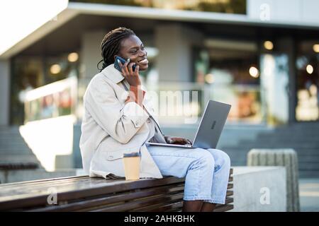 Bella black business donna seduta nella città finanziaria, con computer portatile avente chiamata conversazione, sorridente all'esterno. Professional donna wo Foto Stock