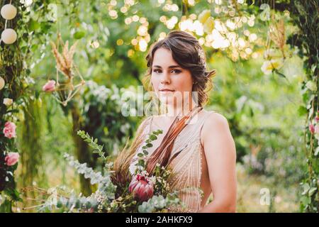 Bella Sposa con bouquet di fiori si erge su sfondo di foresta. Stile rustico. bella sposa abito delicato all'esterno. Close up ritratto di Giovane Foto Stock