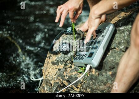 Maschio caucasico di eseguire musica utilizzando il campionatore di natura seduto su una roccia nei pressi delle rive del cristallo limpido fiume. Soleggiato uno scenario rilassante. Foto Stock