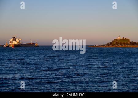 Il Carbone di Bulk Carrier Nave e rimorchiatori partenza dal porto di Newcastle sul pomeriggio soleggiato con Nobbys Capo Faro in background Foto Stock