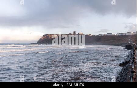 Onde ruggito verso una spiaggia con un terreno non lavorato con una rovina del castello e alberghi nello skyline della citta'. Le ringhiere sono in primo piano. Un cielo blu è sopra. Foto Stock