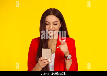 Donna che guarda ad un messaggio nel suo sorriso di telefono e rivolto a voi la fotocamera con il dito. Razza mista modello isolato su sfondo giallo con copia spazio. Foto Stock