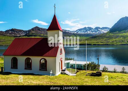 La chiesa del villaggio Mjoifjordur e il paesaggio del fiordo e costo di montagna in background. Austurland, Fjardabyggd, Islanda Orientale Foto Stock