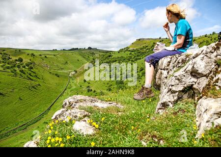 Una donna seduta di mangiare uno spuntino su una roccia che si affaccia Cressbrook Dale Derbyshire Parco Nazionale di Peak District Inghilterra REGNO UNITO Foto Stock