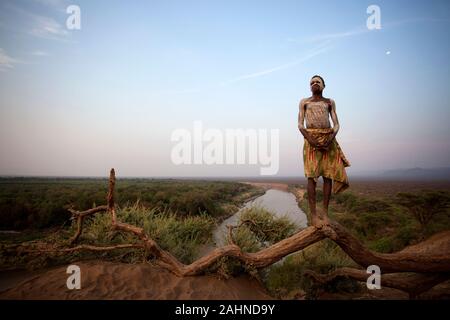 Karo tribù, giovani tribal Karo ragazzo con tradizionalmente faccia dipinta in piedi su un ramo sopra il fiume Omo, valle dell'Omo, Etiopia, Africa Foto Stock