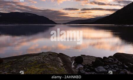 Soluzione satura di Cielo di tramonto riflesso sul ancora, fiordo calmo lago durante un inverno freddo giorno come visto da un'isola nel centro della valle scolpito. Foto Stock