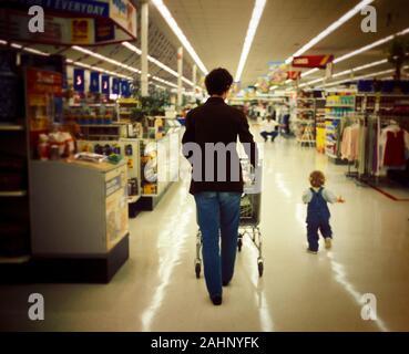 Una donna che spinge un carrello all'interno di un supermercato mentre il suo bambino corre avanti. Foto Stock