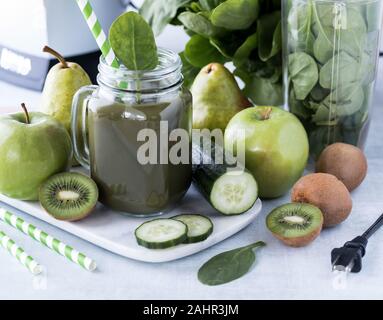 Una chiusura di un vaso di vetro tazza riempita con frullato verde circondato dal verde della frutta e verdura nonché il frullatore. Foto Stock