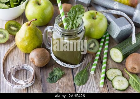 Una vista dall'alto in basso di un barattolo di vetro riempito con il frullato verde circondato dal verde della frutta e verdura e manubri e un nastro di misurazione. La dieta e pesare Foto Stock