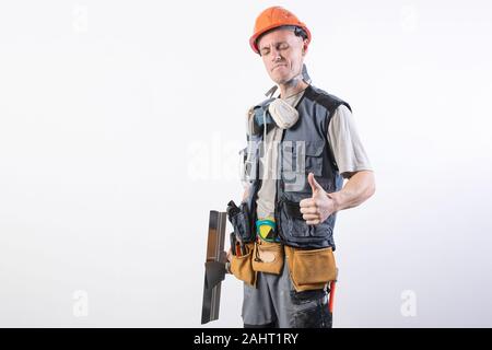 Builder con una spatola. E mostra Thumbs up. In abiti da lavoro e di elmetto. Su uno sfondo grigio chiaro. Foto Stock