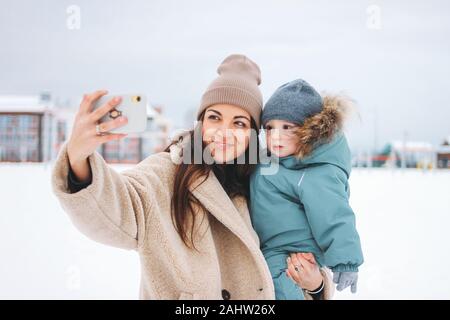 Giovani piuttosto mom brunette donna con simpatici baby boy in tuta calda facendo selfie sulla strada invernale Foto Stock