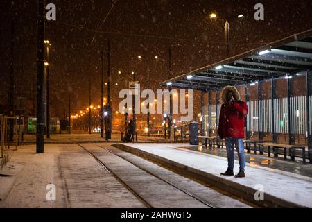 Uomo in rosso cappotto invernale permanente al bus tram fermata in attesa per i mezzi di trasporto pubblico Foto Stock