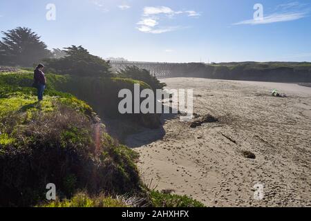 Una donna in un maglione e jeans sorge sulla sommità di basse scogliere coperte di vegetazione verde e si affaccia su una spiaggia di sabbia a Fort Bragg, California. Foto Stock