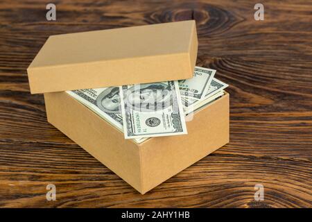 piccola scatola di cartone piena di cento banconote da dollaro primo piano con fuoco selettivo su sfondo di legno naturale Foto Stock