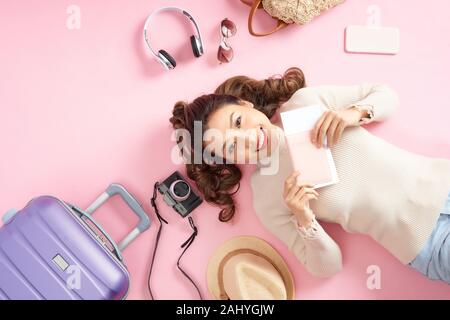 Donna asiatica tenendo il suo passaporto e giacente circondano il suo bagaglio di viaggio sul pavimento di colore rosa. Vista superiore Foto Stock