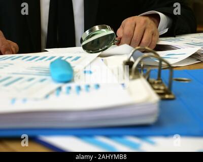 Concetto di audit. Un uomo si siede a un tavolo e controlla i documenti. Pila di carte e di una lente di ingrandimento. Foto Stock