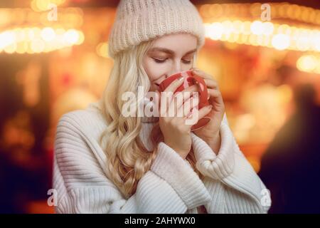 Giovane donna gustando una tazza di caffè caldo in inverno coppettazione nelle sue mani come ella beve con un magico sorriso contro un incandescente caldo arancio backgroun Foto Stock