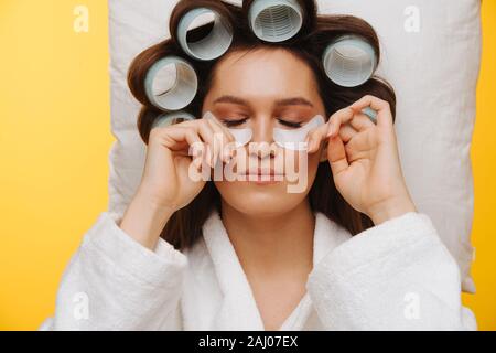 La donna si trova sul cuscino con i capelli in rulli premendo le patch sotto gli occhi per le guance Foto Stock