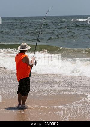 Un uomo sul Fire Island è la pesca nell'oceano indossando un arancione camicia senza maniche e in piedi sul bordo dell'acqua. Foto Stock