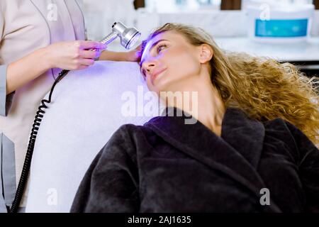 Luce terapia a raggi infrarossi. Testa di Cosmetologia procedura. Close-up di bella donna faccia ricevente a led blu la terapia della luce. Salone estetico dispositivo. Trattamento viso Foto Stock