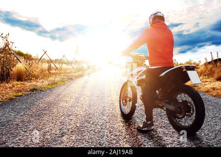 Giovane uomo alla guida con la sua moto su una strada, guardando il tramonto e godendo della libertà e di uno stile di vita attivo. Foto Stock