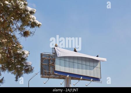 Lavoratore prepara il tabellone per installare il nuovo annuncio. Scalatore industriale di lavorare su una scala - il posizionamento di un banner pubblicitario. Foto Stock