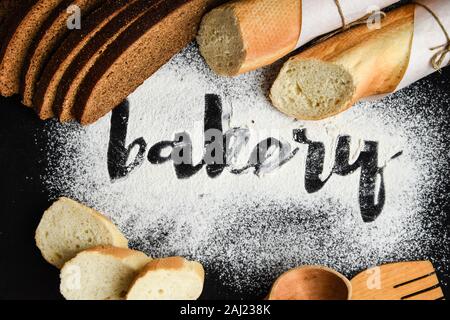 Iscrizione panetteria sulla farina di frumento bianca sparsi affettato baguette francese e il pane di segale con il cucchiaio di legno su sfondo scuro, scritto a mano in farina il Foto Stock