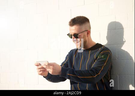 Positivo ragazzo sorridente in informale con il telefono cellulare. Giovane uomo caucasico permanente al outdoor muro bianco e vedere video divertenti nel suo dispositivo. Tempo libero Foto Stock