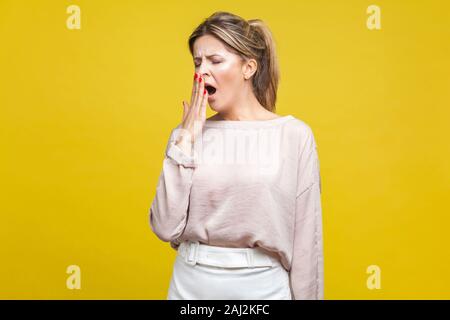 Ritratto di sleepy giovane donna con capelli biondi in casual camicetta beige sbadiglia permanente con gli occhi chiusi e la copertura della bocca, sensazione di esaurimento bisogno di riposo Foto Stock