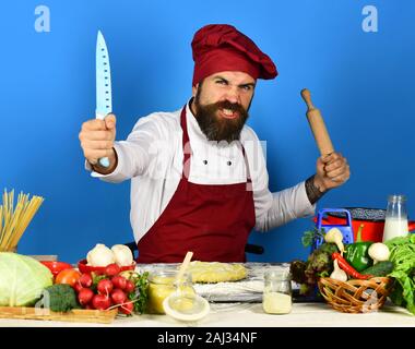 Chef con pasta, verdure e la pasta sul tavolo. Uomo con la barba si siede da bancone su sfondo blu. Il processo di cottura del concetto. Cucinare con il viso arrabbiato in Borgogna mantiene uniforme matterello e coltello Foto Stock