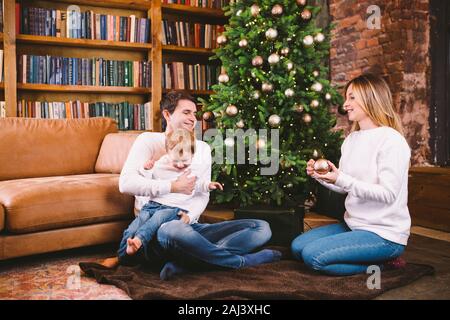 Natale in famiglia concetto. Bella famiglia siede vicino a albero di Natale di sera d'inverno. La famiglia felice con il figlio sul pavimento vicino al divano di casa. La famiglia a casa Foto Stock